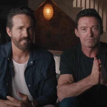 Ryan Reynolds and Hugh Jackman are sitting next to each other on a couch talking to the camera.
